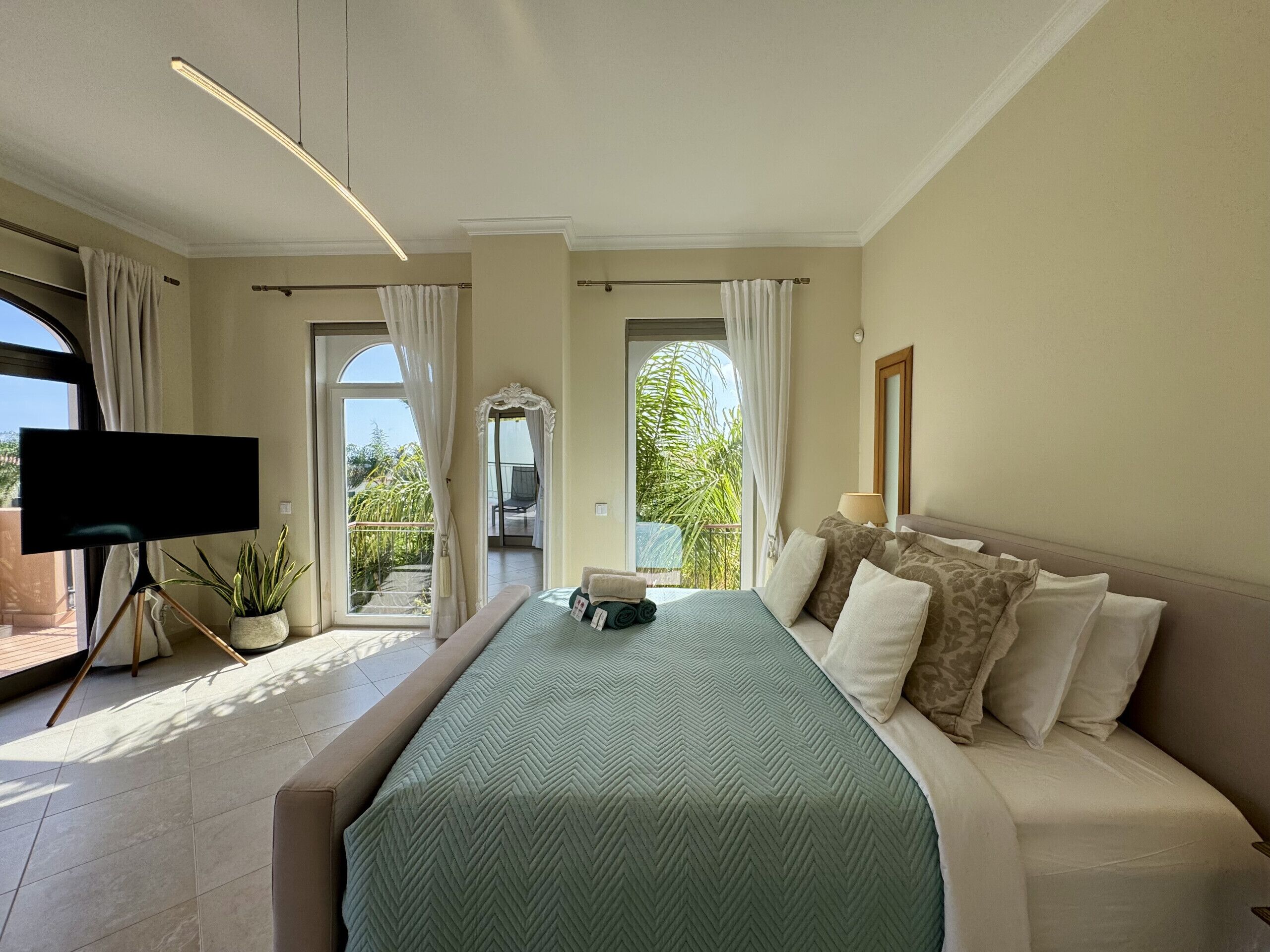 A hotel room with a large bed, a TV, a balcony with curtains, and a view of greenery.