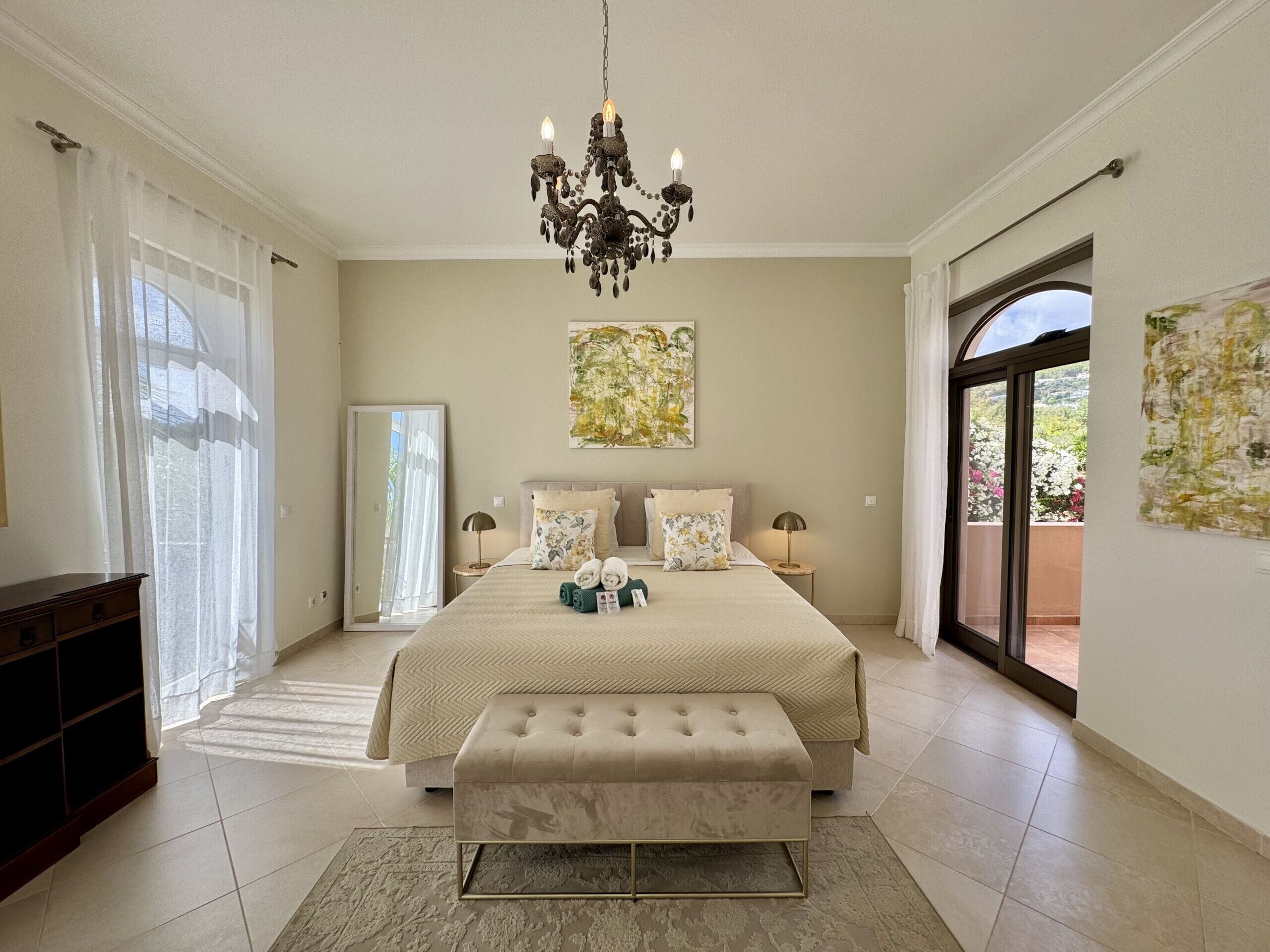 A bedroom with a large bed, a bench, a chandelier, and a view of the outdoors.