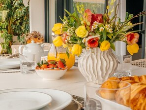 Ein Frühstücksbuffet mit Blumen in einer Vase, Obstschale, Orangensaftkaraffe und Croissants auf einem Tisch.
