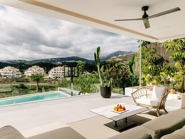 Ein moderner Außenbereich mit Sitzgelegenheit, einem Pool, einem Tisch mit Früchten und Blick auf Gebäude und Berge.