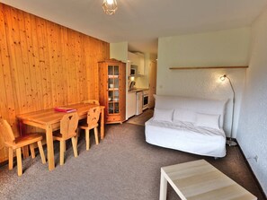Ein gemütlicher Raum mit Holztisch, Stühlen, einem weißen Sofa, einem Schrank mit Glastüren und einer kleinen Küchenzeile.