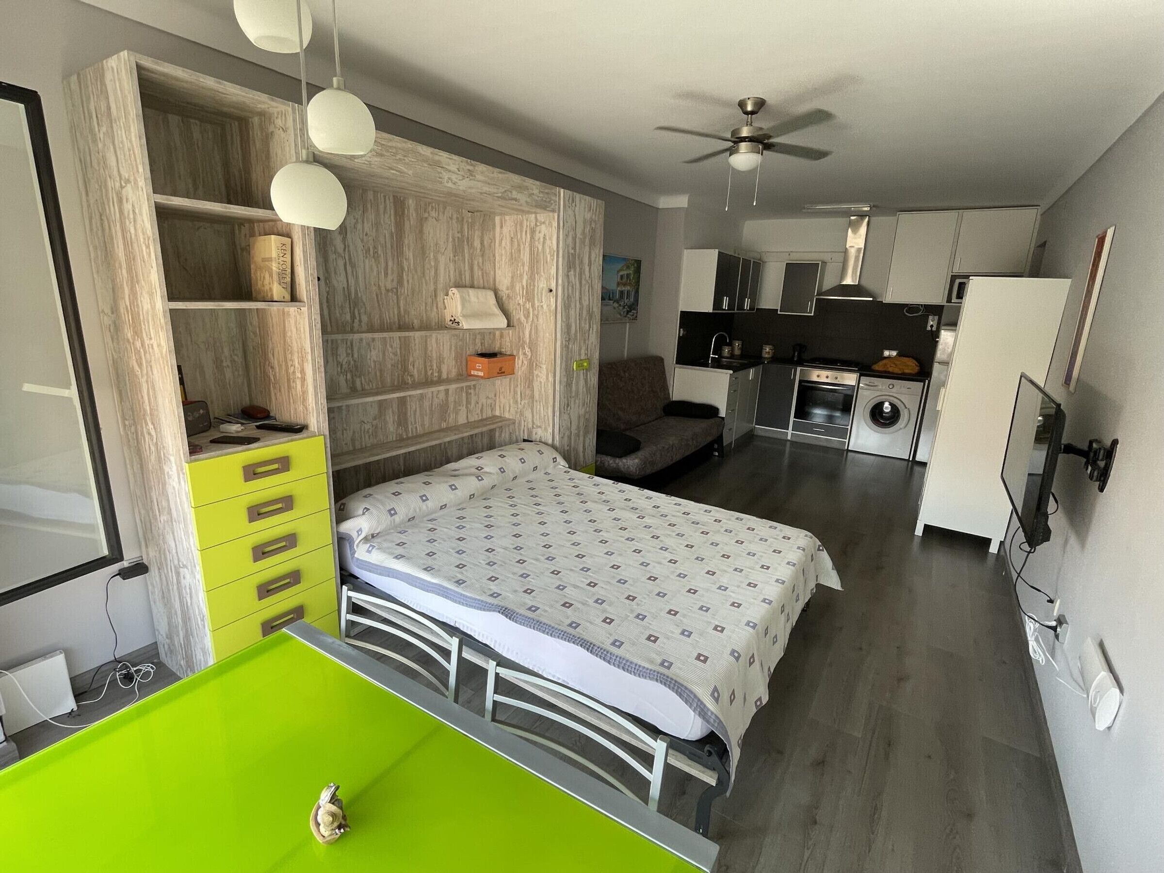 A compact bedroom with a bed, a green desk, and a wardrobe. A kitchenette with a stove, sink, and refrigerator is visible in the background.