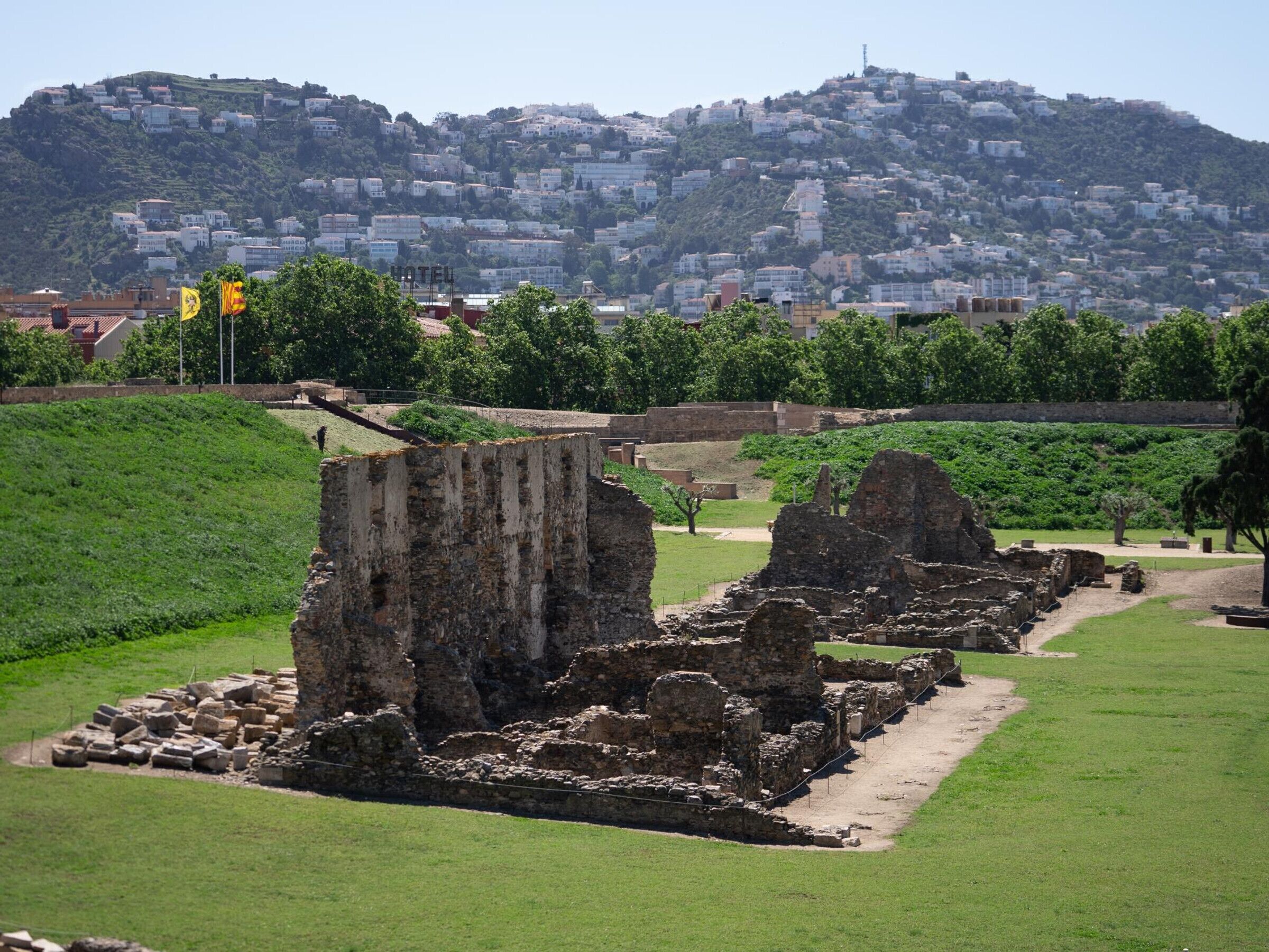Ruinas antiguas con muros de piedra y piedras dispersas, frente a una colina con edificios.