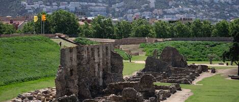 Ruinas antiguas con muros de piedra y piedras dispersas, frente a una colina con edificios.