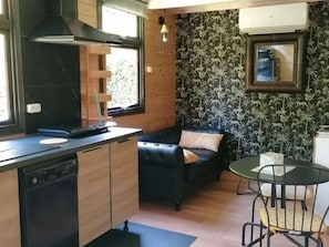 A compact kitchen with a dishwasher, a built-in stove, and a window. There is a black sofa with patterned cushions and a small dining table with chairs. - Cozy chalet with AC - 2 guests (Noves)