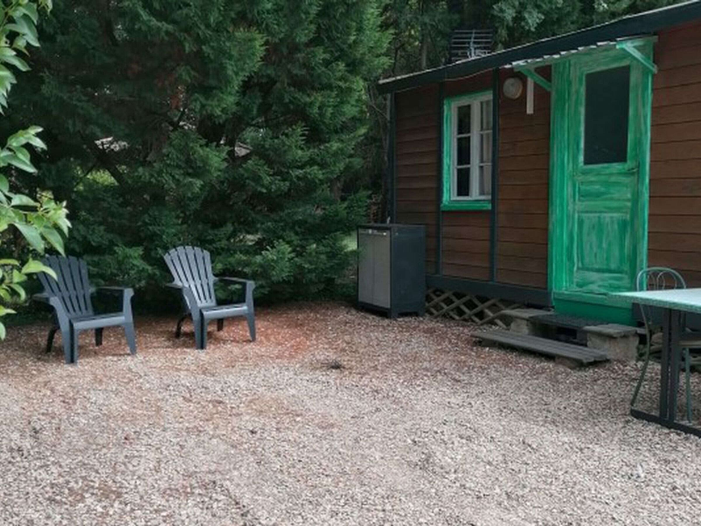 Une cabane avec un sol recouvert de gravier, deux fauteuils Adirondack, une table et une porte verte.