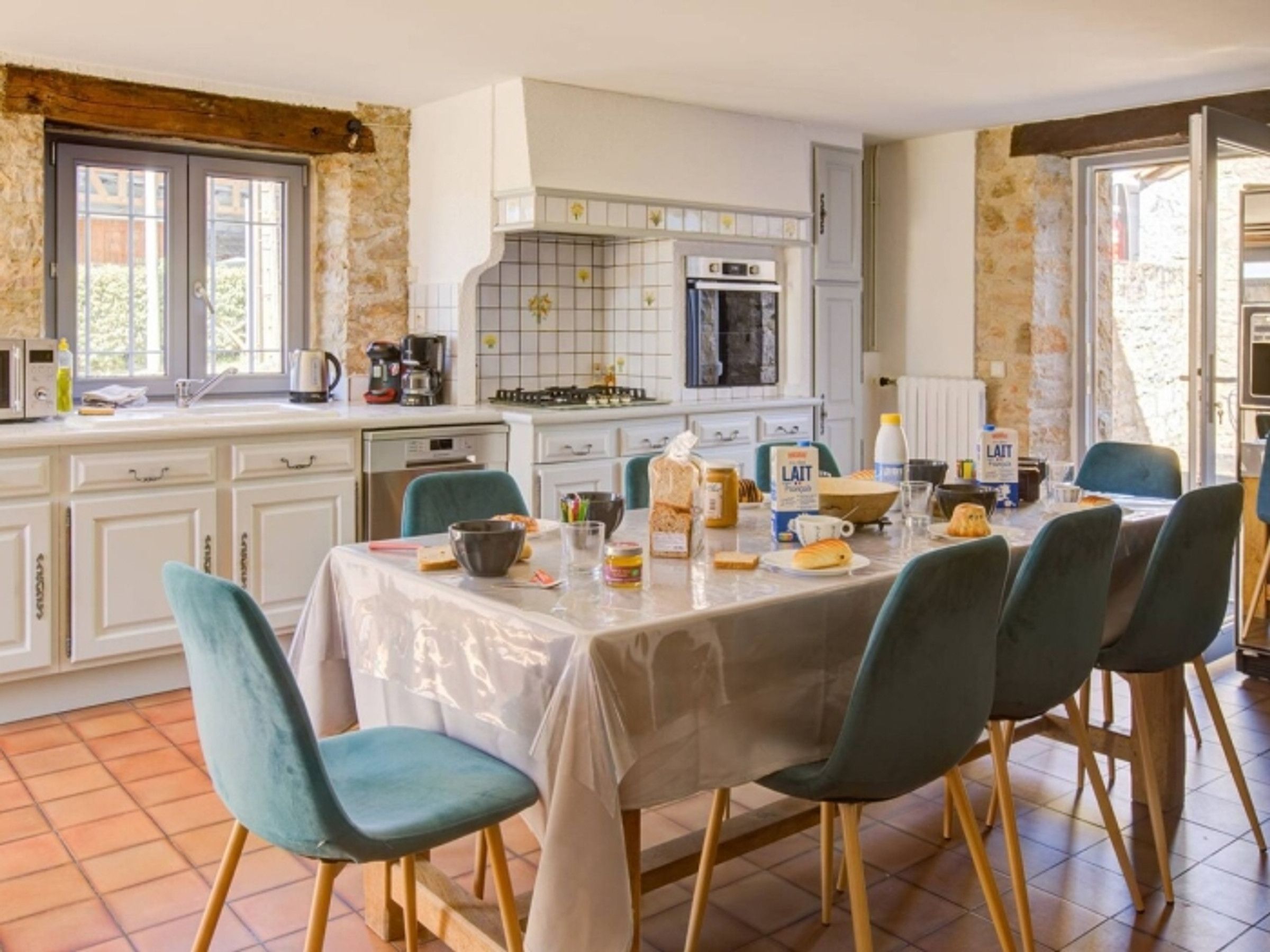 On aperçoit un coin repas avec une table dressée pour quatre personnes, entourée de chaises. Au fond, on distingue une cuisine équipée d’une cuisinière, d’un four et d’un évier.