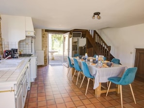 Une cuisine spacieuse avec un coin repas, comprenant une table dressée pour six personnes avec des chaises bleues, un escalier et une vue sur l’extérieur.