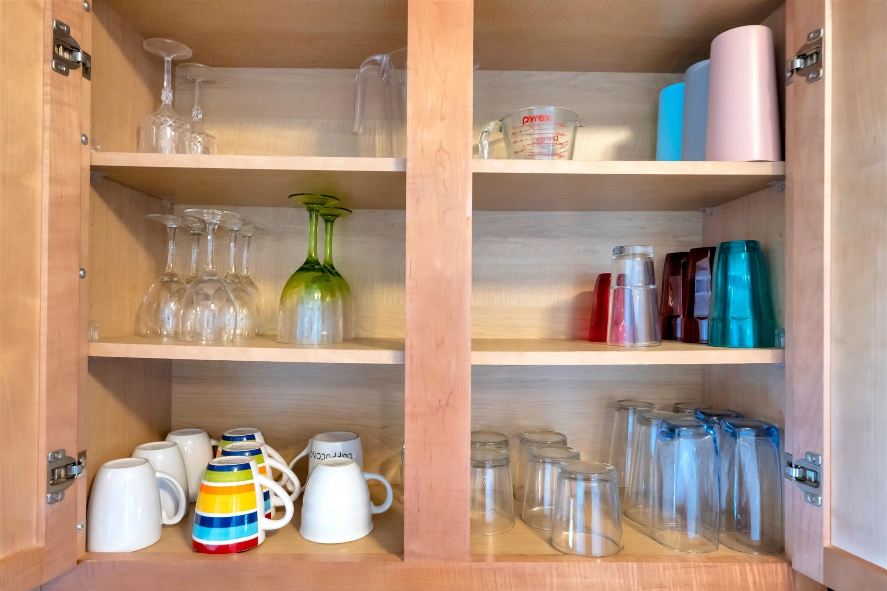 A wooden cabinet with shelves containing various glassware, including wine glasses, a green vase, and colorful mugs.
