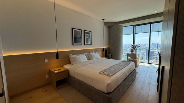 Une chambre moderne avec un grand lit, des tables de chevet, une vue sur la ville et des œuvres d’art encadrées accrochées au mur.