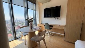 Ein modernes Zimmer mit einem runden Tisch, zwei Stühlen, einem an der Wand montierten Fernseher und einem großen Fenster mit Blick auf die Stadt.