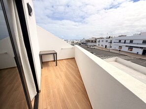 Ein Balkon mit Holzboden, einem kleinen Tisch und Blick auf eine Straße mit Gebäuden und blauem Himmel.