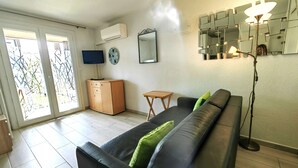 A living room with a black leather sofa, a wooden cabinet, a television, a wall clock, a mirror, and a floor lamp. - Studio cabin, secure parking - 4 guests (Cap d'Agde)
