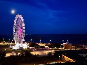 Ein Riesenrad bei Nacht, mit einem Mond am Himmel, umgeben von einer Parkanlage mit Wegen und einem kleinen Gebäude.