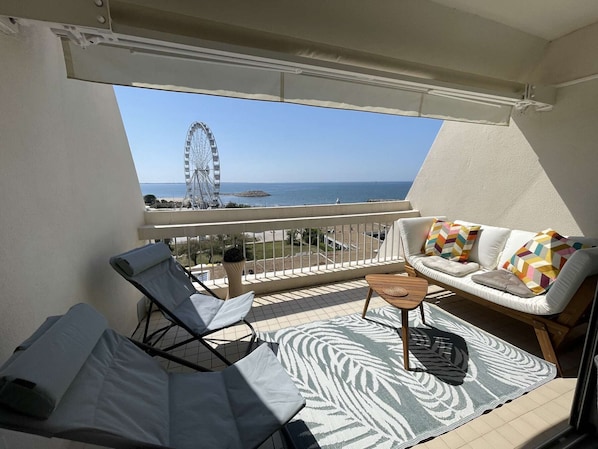 Ein Balkon mit Blick auf das Meer und ein Riesenrad, möbliert mit einer Couch und zwei Liegestühlen.