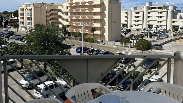 Ein Blick auf ein mehrstöckiges Gebäude mit einer geometrischen Fassade, einen Parkplatz mit Autos und eine Straße mit Fußgängern.