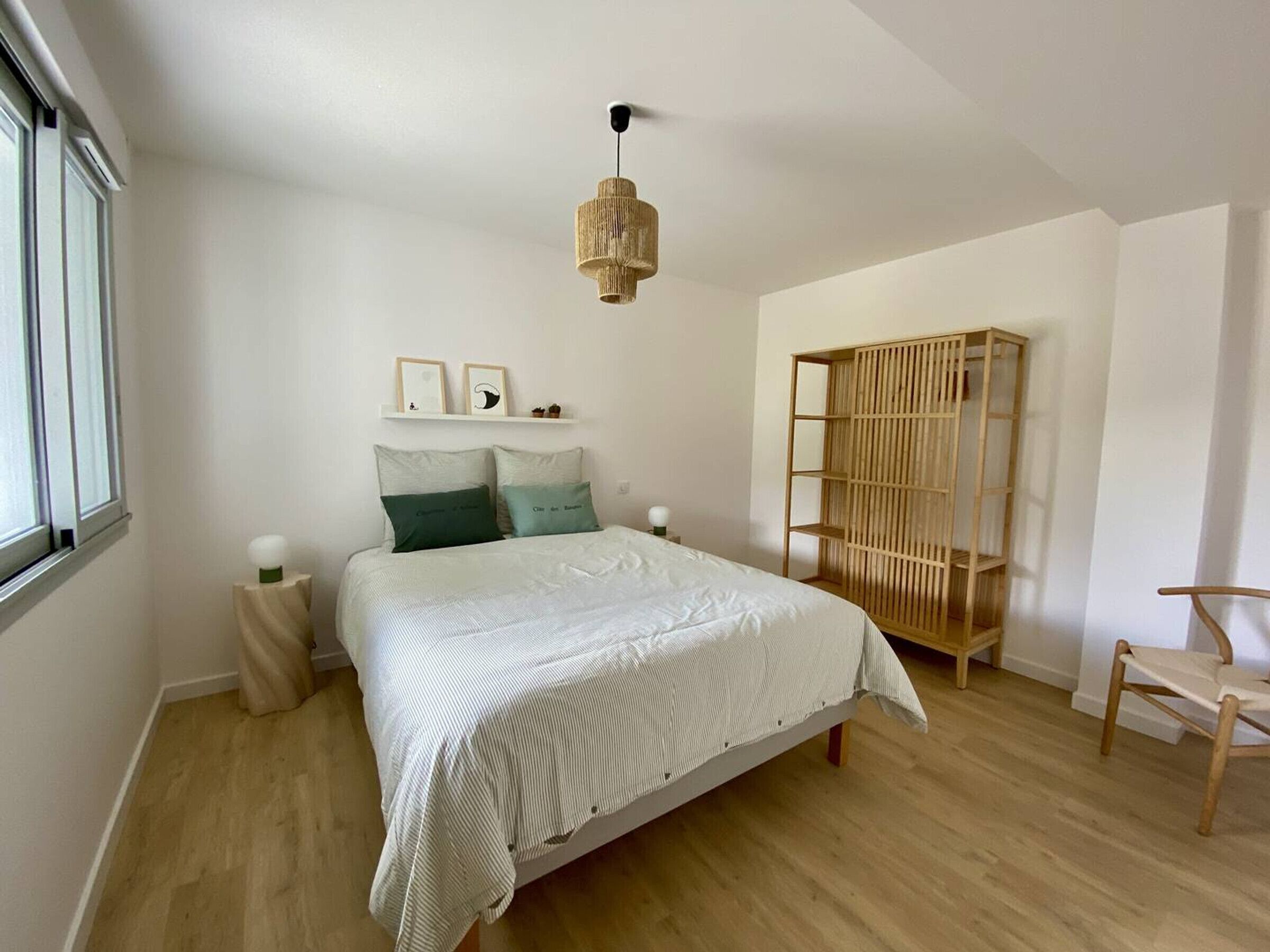 A bedroom with a bed, a bedside table, a hanging light fixture, a wooden wardrobe, and a chair.
