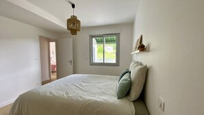 Een slaapkamer met een bed, een raam, een hanglamp en een deur naar een andere kamer.