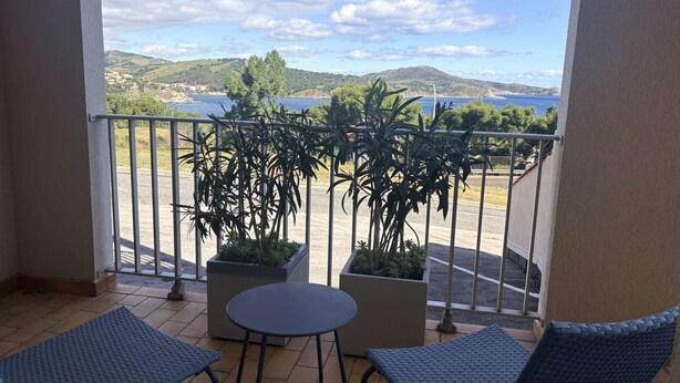 Un balcone con due sedie e un tavolo, affaccia su un'area di parcheggio e un panorama che include colline e un tratto d'acqua.