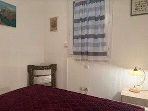 Ein Schlafzimmer mit Bett, Nachttischlampe, einem Fenster mit Vorhängen, einem gerahmten Bild und einem an der Wand montierten Lichtschalter.