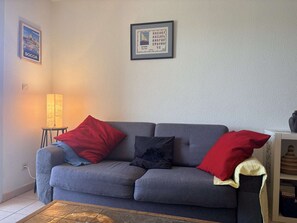 Ein graues Sofa mit roten und schwarzen Kissen, ein kleiner Tisch mit einer Lampe und ein gerahmtes Bild an der Wand.
