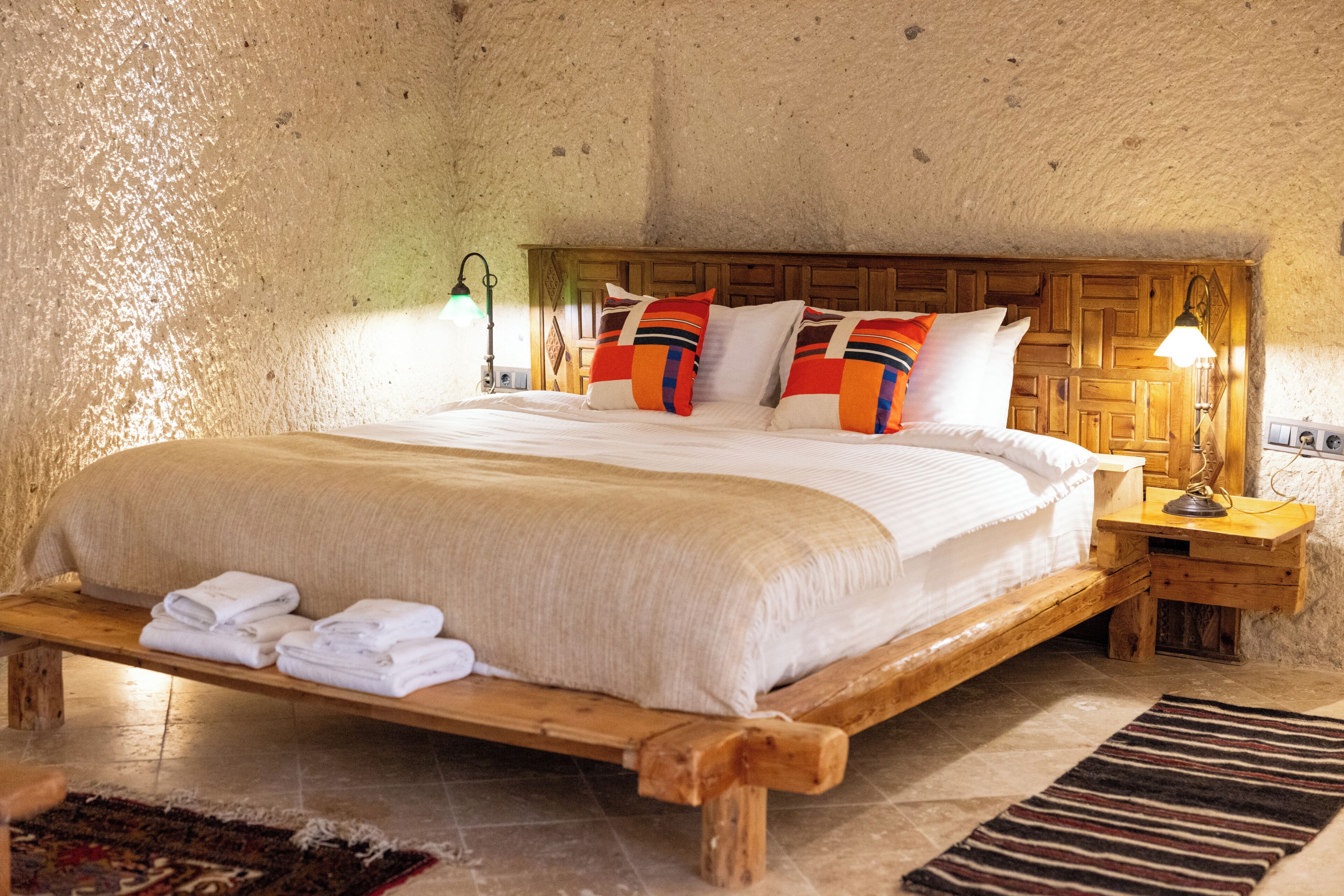 Un dormitorio rústico con una cama de madera, almohadas, un banco con toallas y una mesita de noche con lámpara.