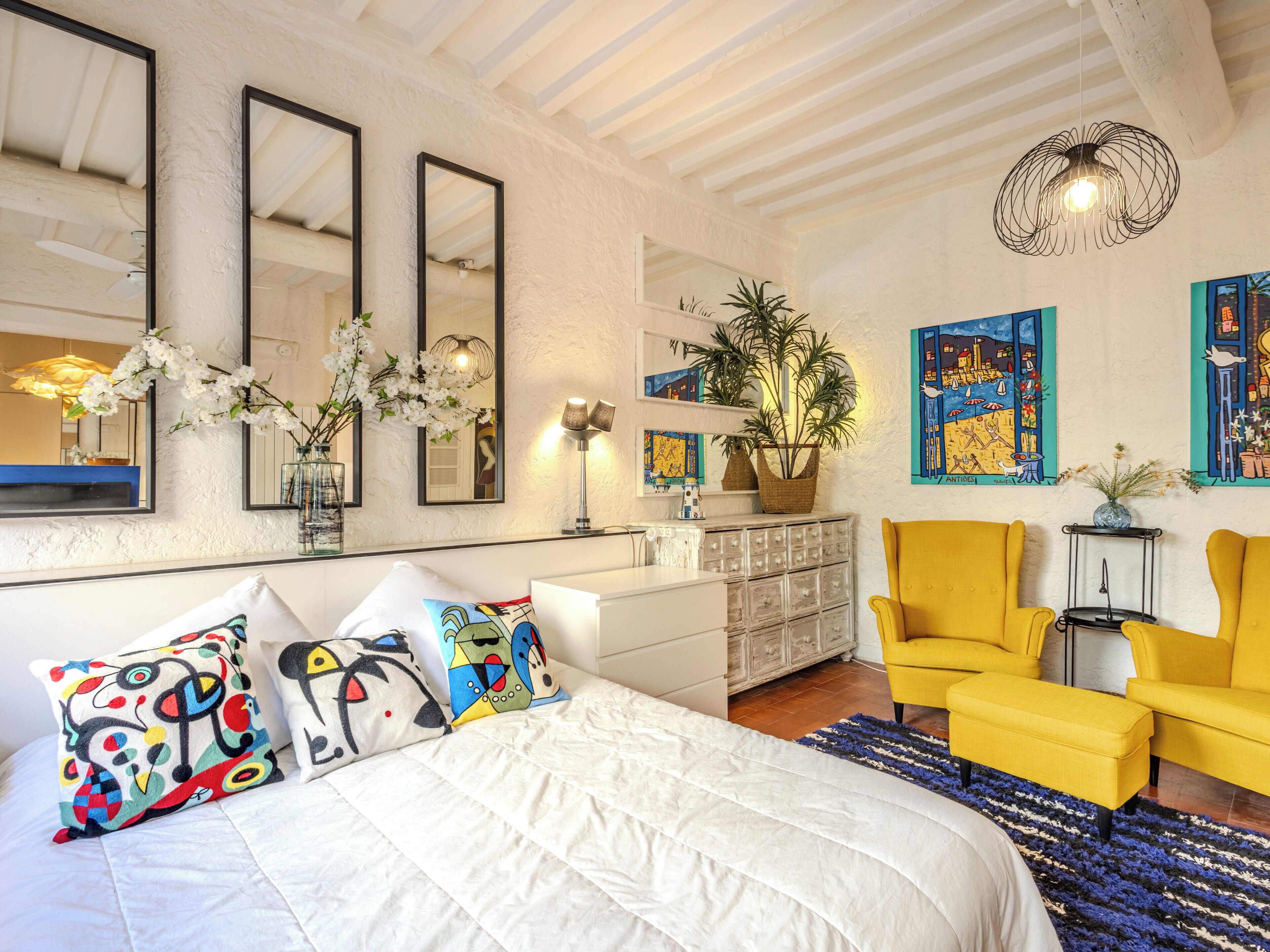 A bedroom with a bed, two chairs, a dresser, a mirror, and wall art.