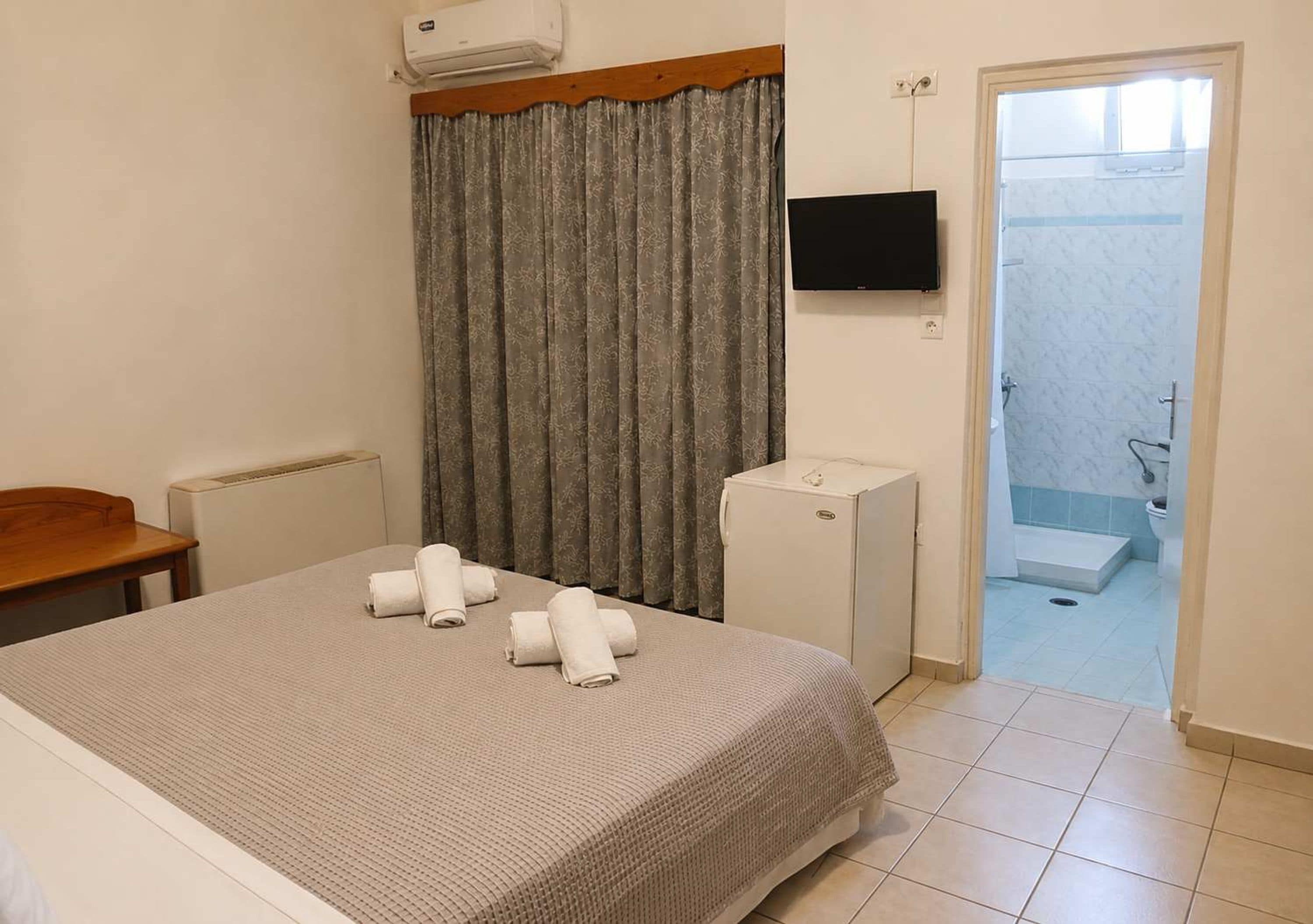Habitación de hotel con cama, escritorio, televisor, nevera pequeña y baño visible a través de una puerta abierta.