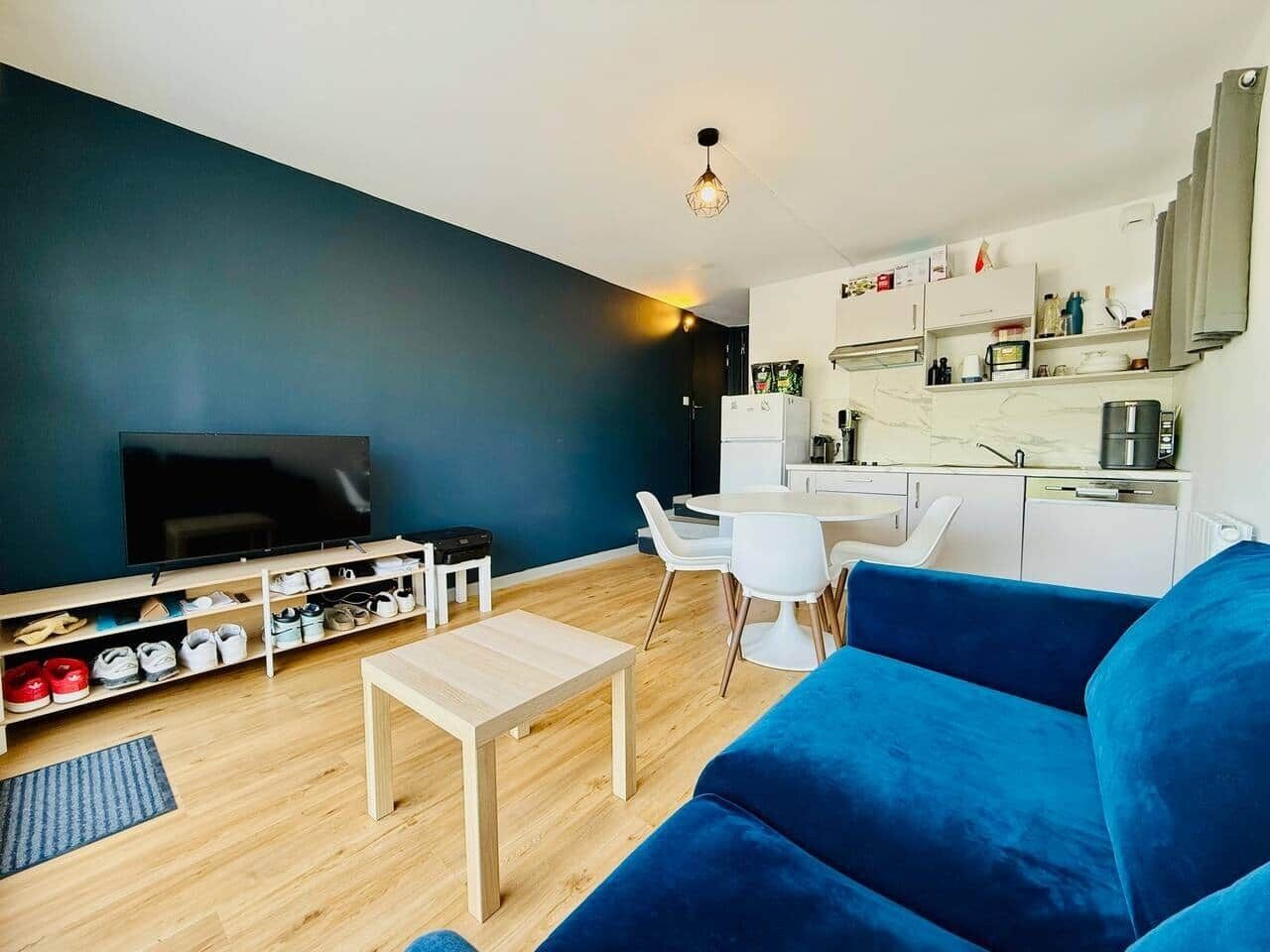 A living room with a blue sofa, a TV, a wooden coffee table, a dining area with a white table and chairs, and a kitchen area with cabinets and appliances.