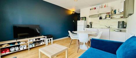 A living room with a blue sofa, a TV, a wooden coffee table, a dining area with a white table and chairs, and a kitchen area with cabinets and appliances.