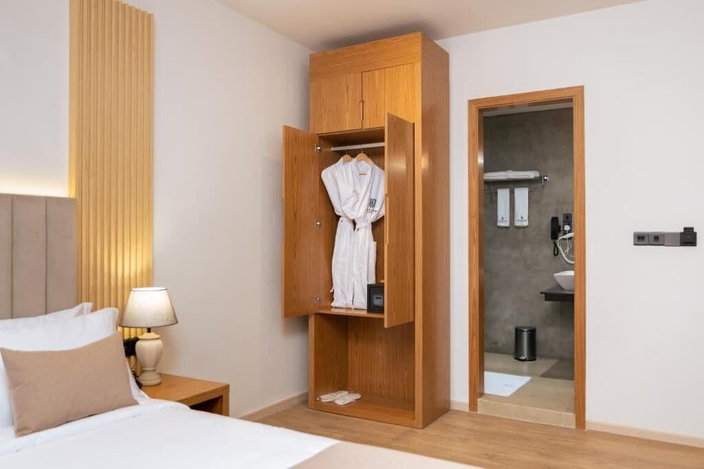Ein Hotelzimmer mit Bett, Nachttisch mit Lampe, einem Holzschrank mit aufgehängtem Bademantel und einem durch eine geöffnete Tür sichtbaren Badezimmer.