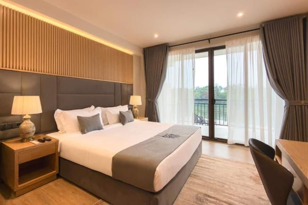 A hotel room with a large bed, two bedside tables with lamps, a chair, a desk, and a balcony with curtains.