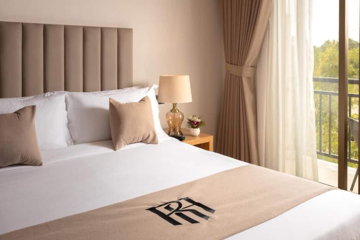 A hotel room with a bed, a nightstand with a lamp, and a view of the outdoors.