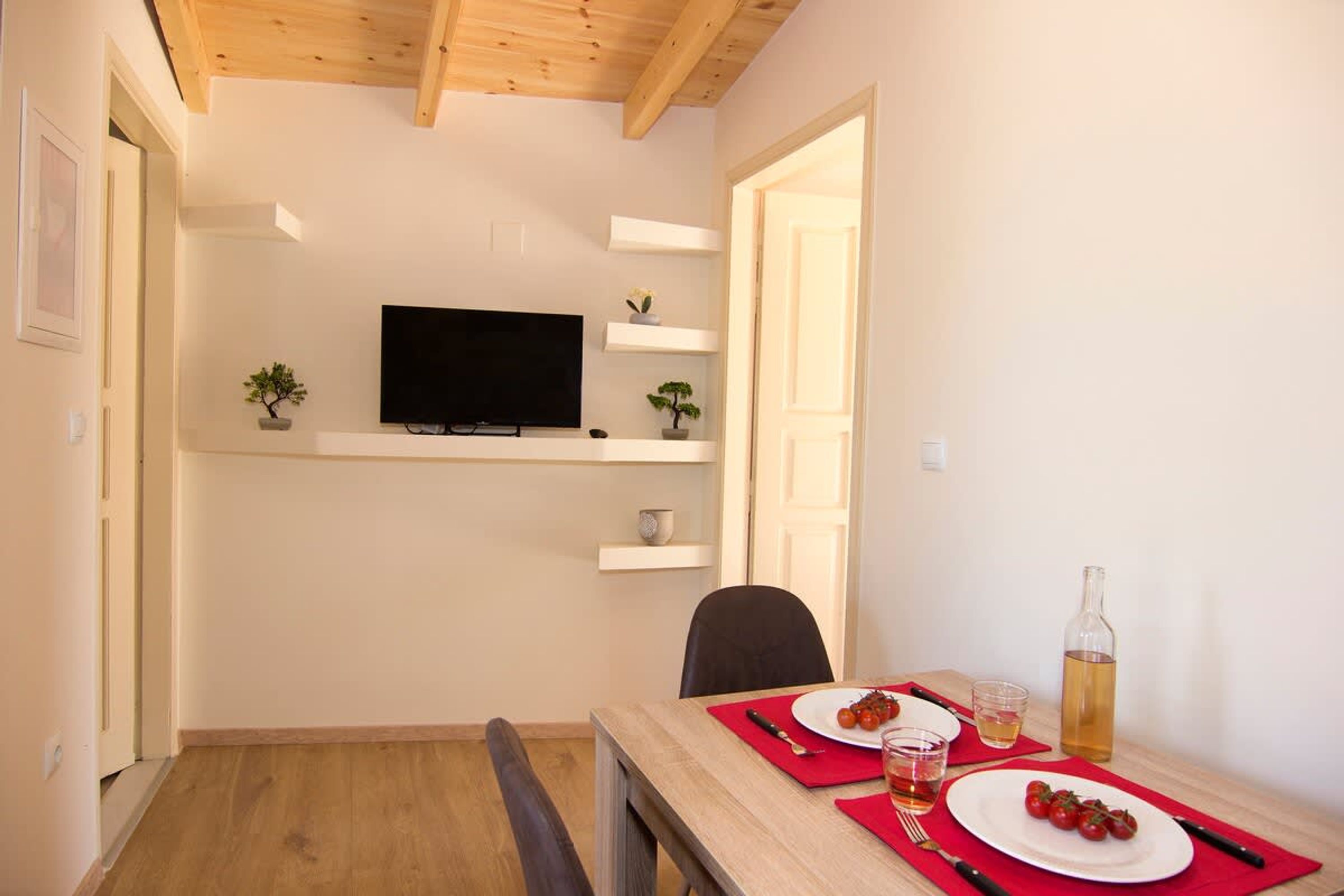 Uma sala de jantar com uma mesa posta para dois, uma televisão em uma prateleira e uma porta que dá para outro quarto.