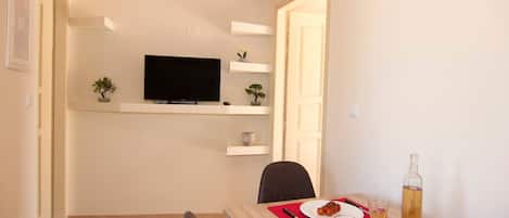 Uma sala de jantar com uma mesa posta para dois, uma televisão em uma prateleira e uma porta que dá para outro quarto.