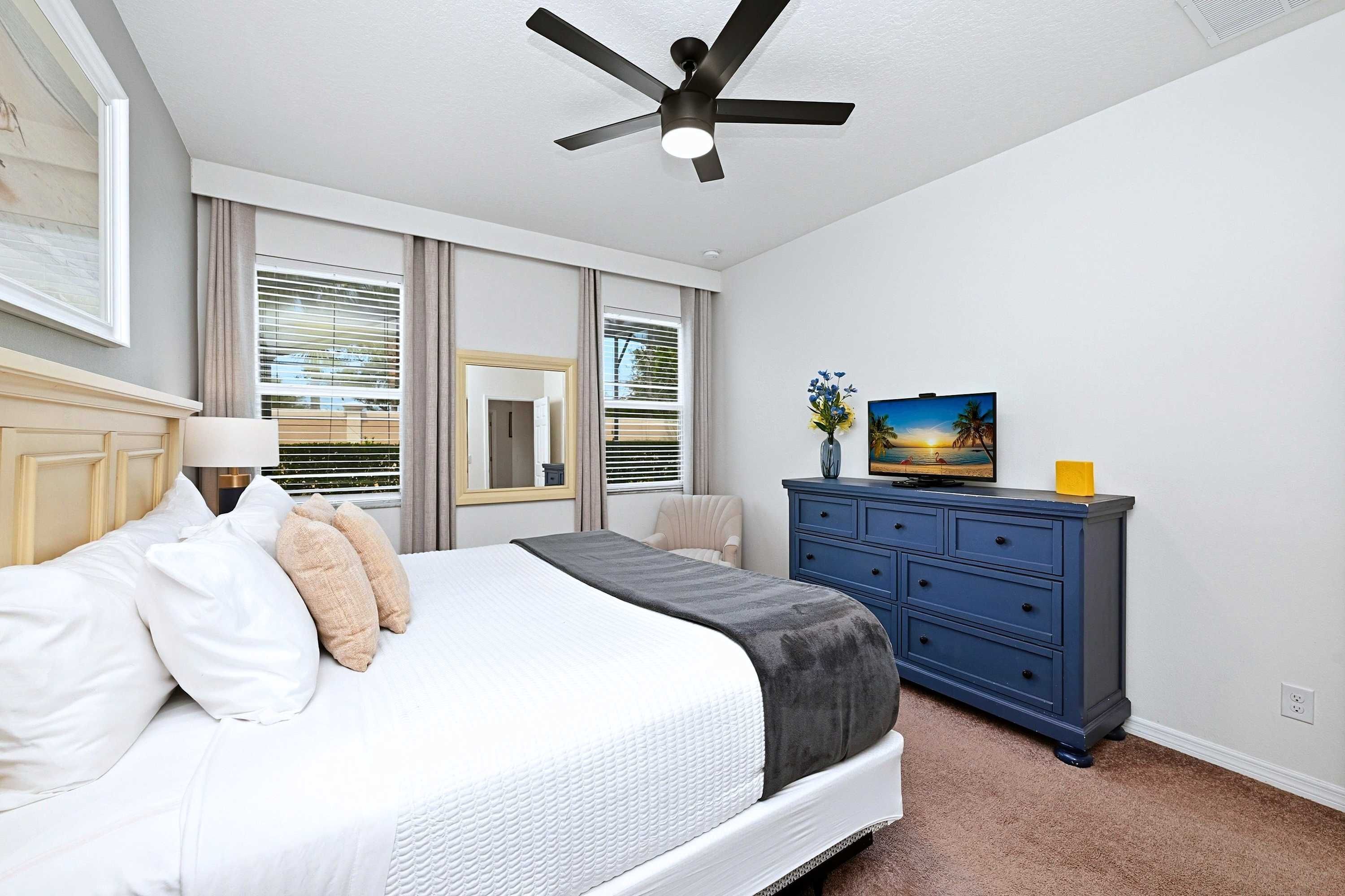 A bedroom with a bed, a ceiling fan, a dresser, a television, and a window with blinds.
