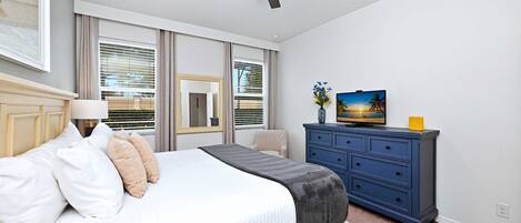 A bedroom with a bed, a ceiling fan, a dresser, a television, and a window with blinds.