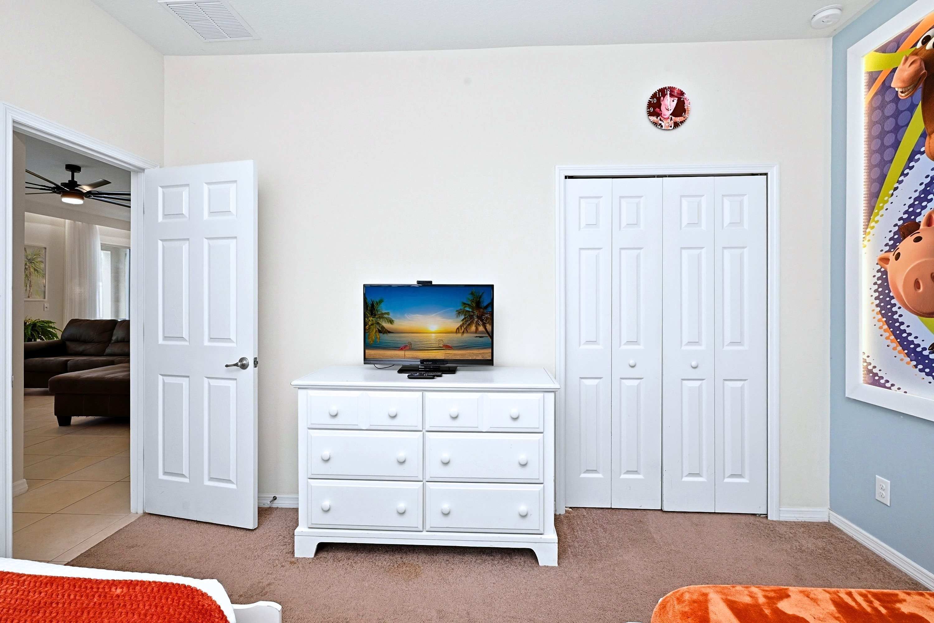 A bedroom with a TV on a dresser, two white doors, a ceiling fan, and a painting on the wall.