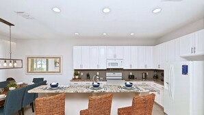 A modern kitchen with white cabinets, a central island with bar stools, a dining area with blue chairs, and a large window. - Upstay Disney Getaway With Pool Game Room (Davenport)