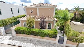 A two-story building with a tiled roof, a gated entrance, and a landscaped garden with various plants. - ALGARVE STUNNING VILLA WITH POOL by HOMING (Santa Bárbara de Nexe)
