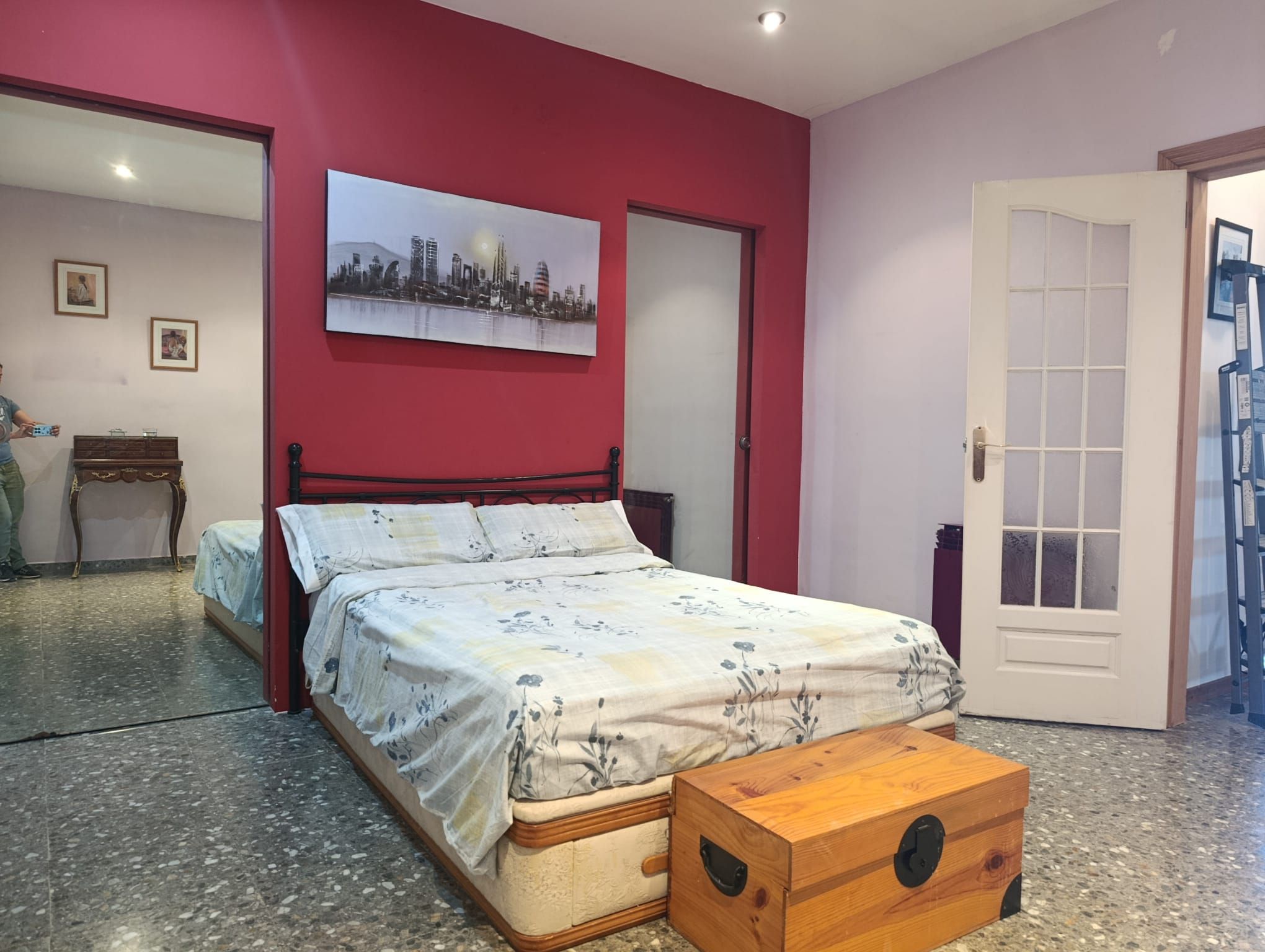A bedroom with a bed, a wooden chest, a cityscape painting, and a door.