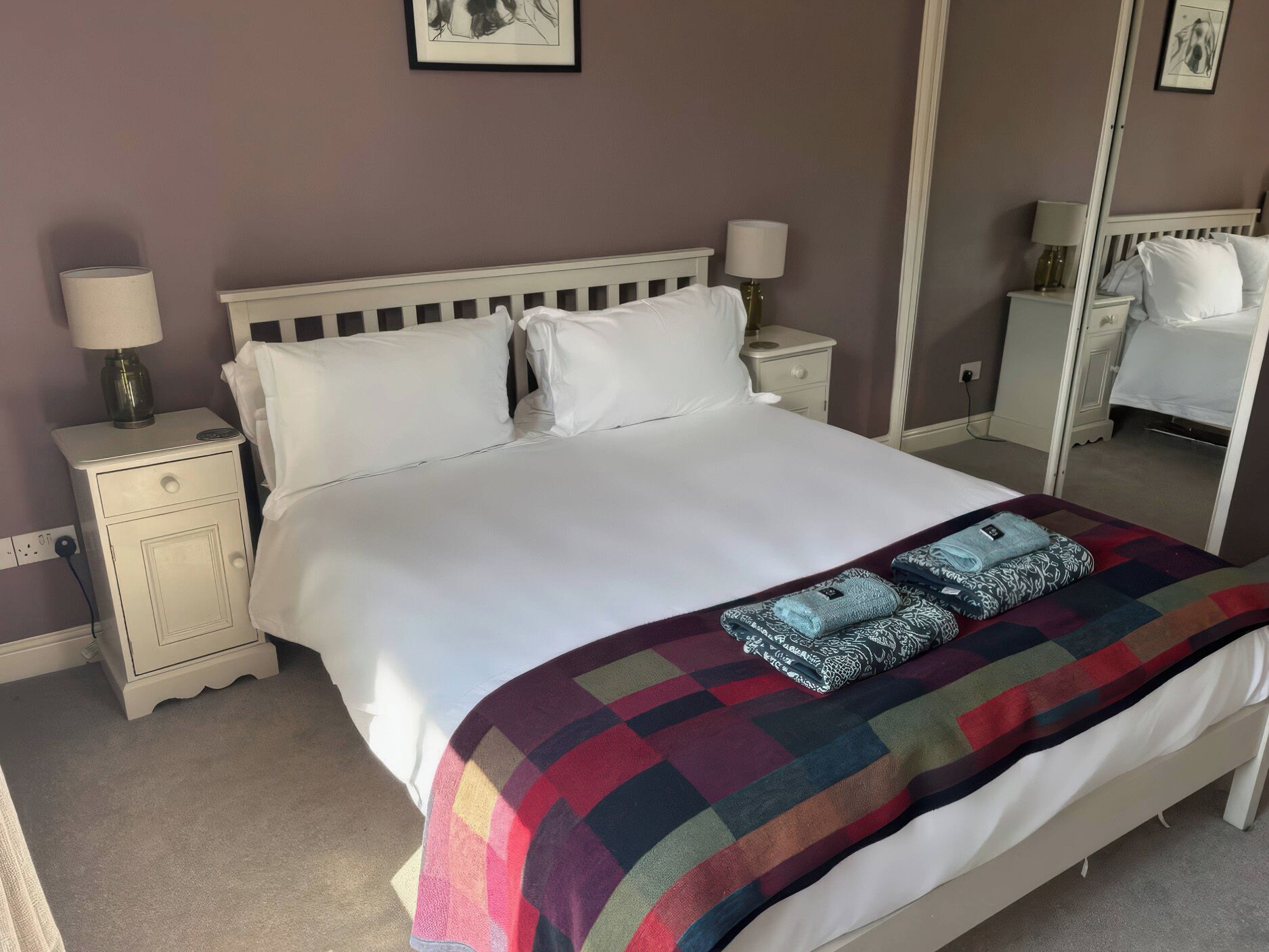A neatly made bed with a colorful blanket and pillows, a bedside table with a lamp, and a mirror on the wall.