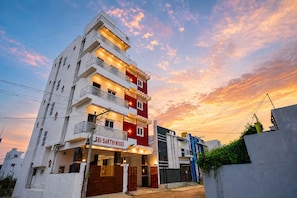 A multi-story building with balconies, a sign reading 'SRI SAKTHINIVAS,' and a sunset sky.