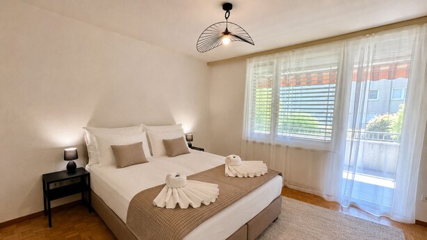 A bedroom with a bed, bedside tables, a ceiling fan, a window with blinds, and a view of buildings outside.