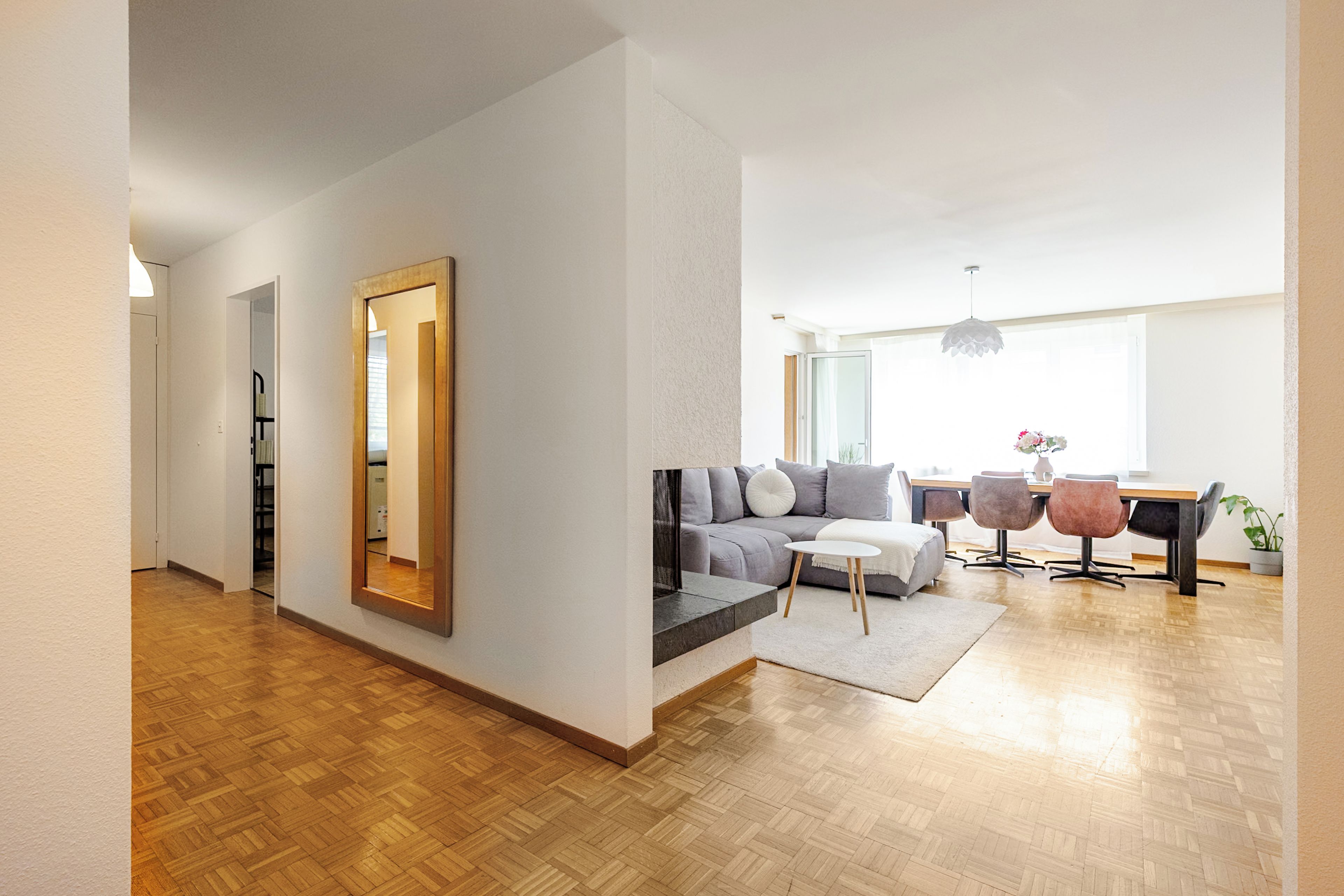 A modern living room with a sofa, coffee table, and chairs, a dining area with a table and chairs, and a large mirror on the wall.
