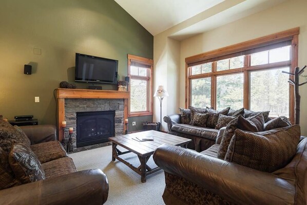 A living room with a stone fireplace, leather sofas, a television, and large windows. - Golf Course Property, Allows Pets, BBQ, Underground Parking, Laundry (Mammoth Lakes)
