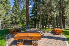 A park with a central stone fire pit, surrounded by benches and tall pine trees. - Golf Course Property, Allows Pets, BBQ, Underground Parking, Laundry (Mammoth Lakes)