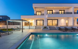 Ein modernes, zweistöckiges Haus mit Swimmingpool, Sitzgelegenheiten im Freien und einer überdachten Terrasse.