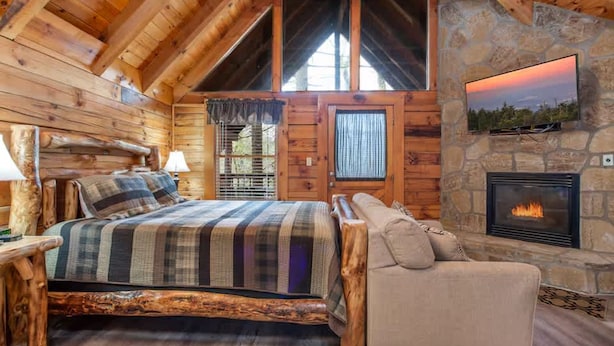 A log cabin bedroom with a bed, a stone fireplace, and a television mounted on the wall. - Location Hot Tub Privacy Fireplace Common Pool (Gatlinburg)