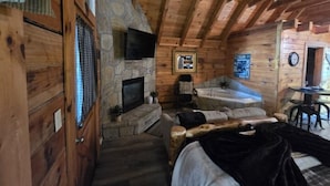 A log cabin interior with a stone fireplace, a stone bathtub, a bed, and a television mounted on the wall. - Location Hot Tub Privacy Fireplace Common Pool (Gatlinburg)